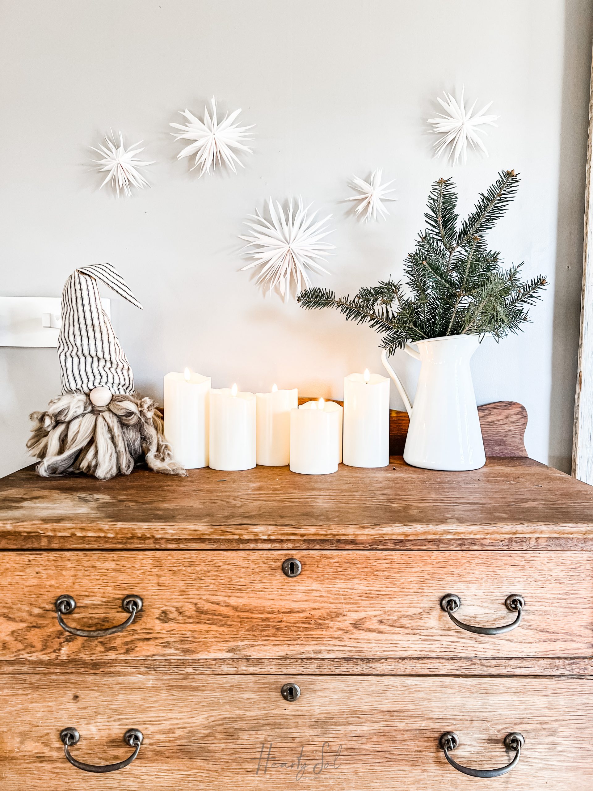 wooden dresser with candles, gnome, white pitcher with greenery in it and 5 white stars on the wall